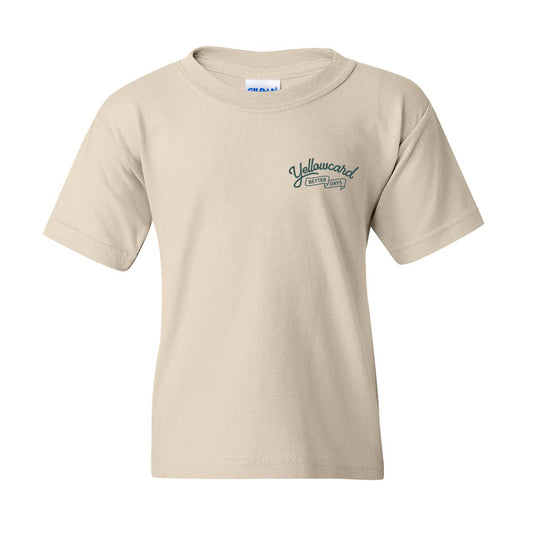 The Better Days Airlines - Natural Youth T-Shirt (Pre-Order) is a 100% cotton, classic fit youth tee in beige, featuring a small dark green Yellowcard Official Crew logo on the left chest, shown against a white background.
